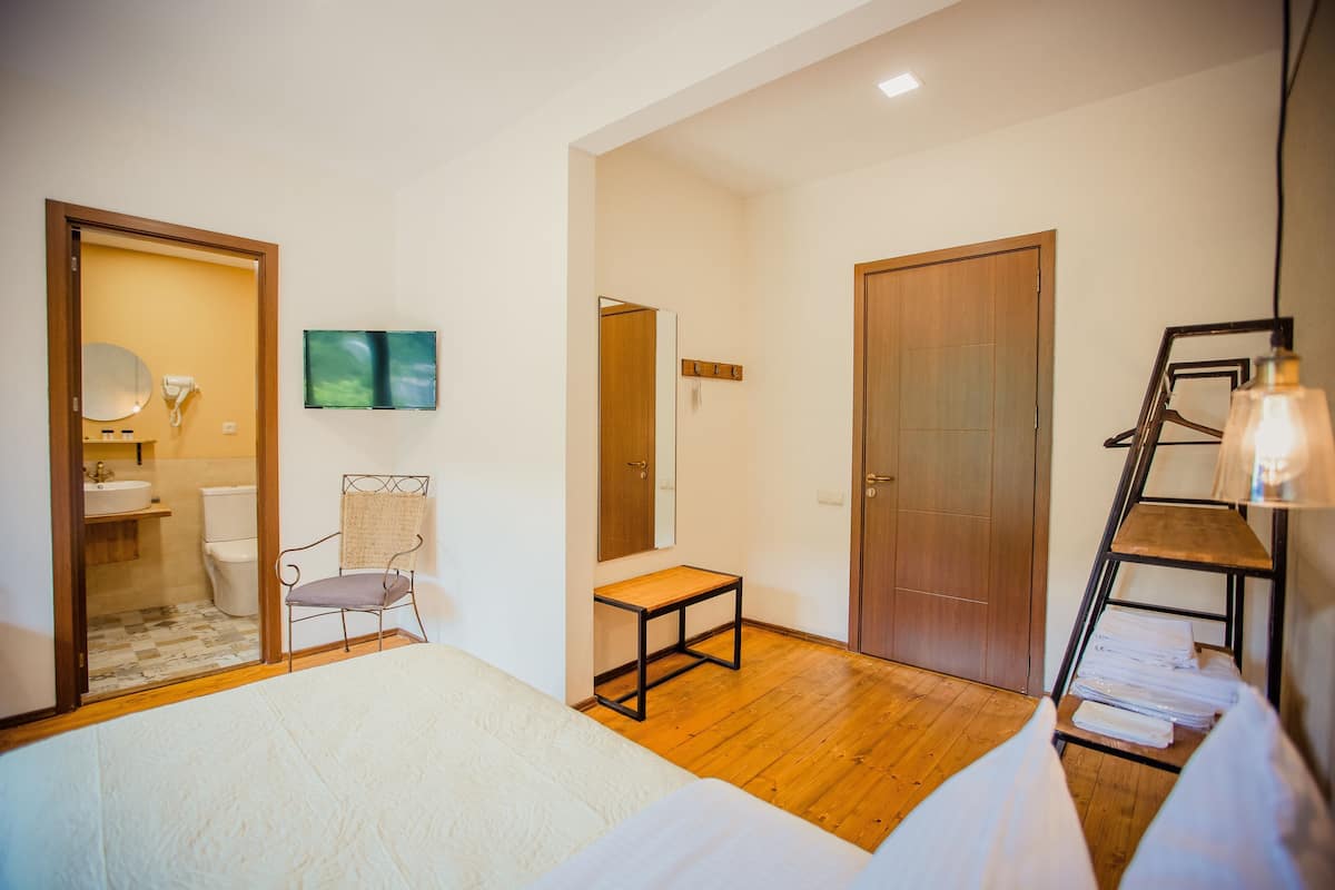 A bedroom with a bed, a wooden floor, a chair, a small table, a mirror, a TV, and a wardrobe.