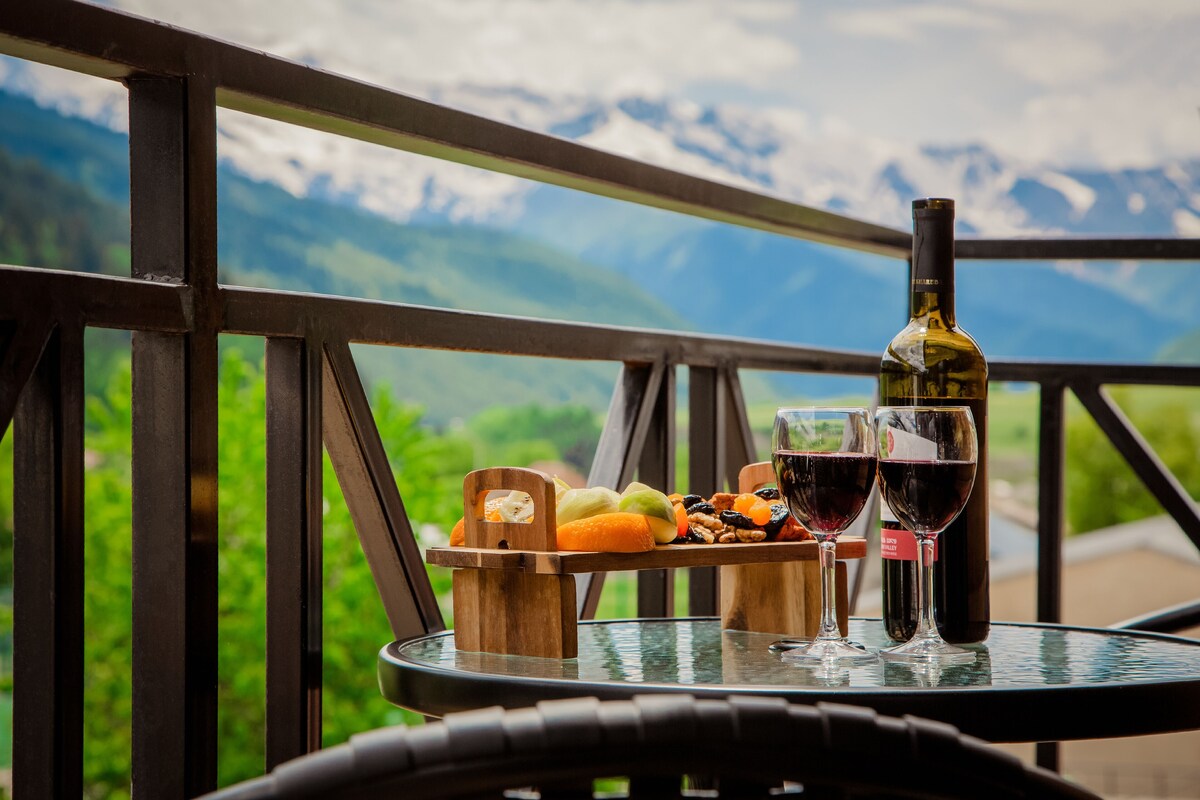 Una mesa con una botella de vino, dos copas y una bandeja de frutas en un balcón con vista a la montaña.
