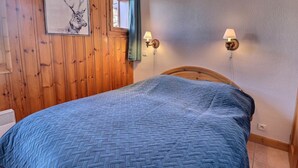 A bedroom with a wooden paneled wall, a bed with a blue quilt, a window with a curtain, and wall-mounted lamps. - Charming 2-room cabin in quiet residence - WiFi included (Méribel)