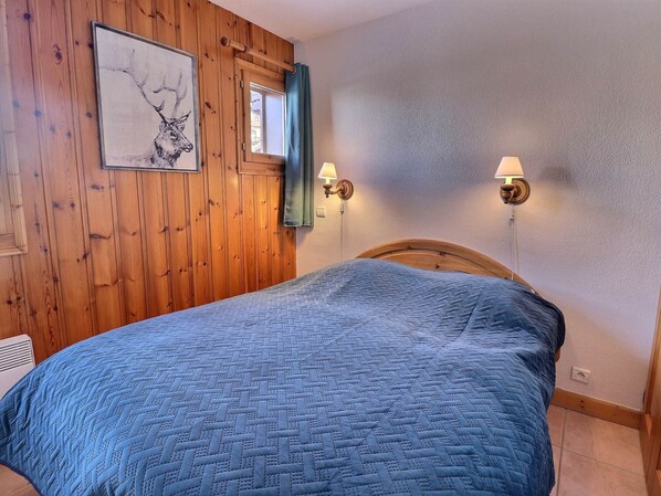 A bedroom with a wooden paneled wall, a bed with a blue quilt, a window with a curtain, and wall-mounted lamps.