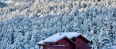 A multi-story building with a red facade, surrounded by snow-covered trees and mountains in the background.