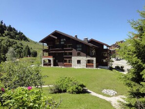 Traditional chalet-style building with a central entrance and multiple balconies, set amidst a well-maintained landscape with grassy areas and flowering plants.