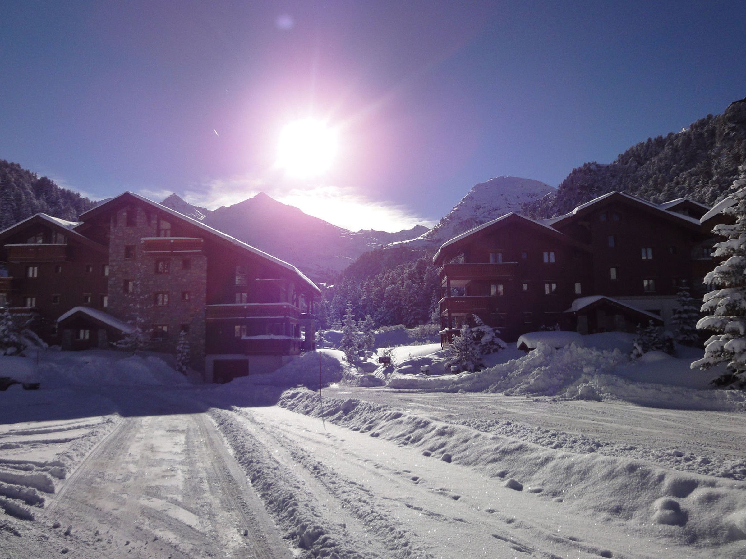 A snow-covered mountain lodge with multiple buildings, a clear sky, and a bright sun.