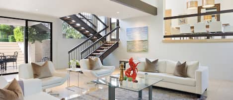 A spacious living room with a modern staircase, a glass coffee table, and a large painting on the wall.