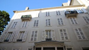 Un bâtiment doté de nombreuses fenêtres et volets, d’une entrée centrale avec une porte décorative et de balcons pourvus de balustrades.