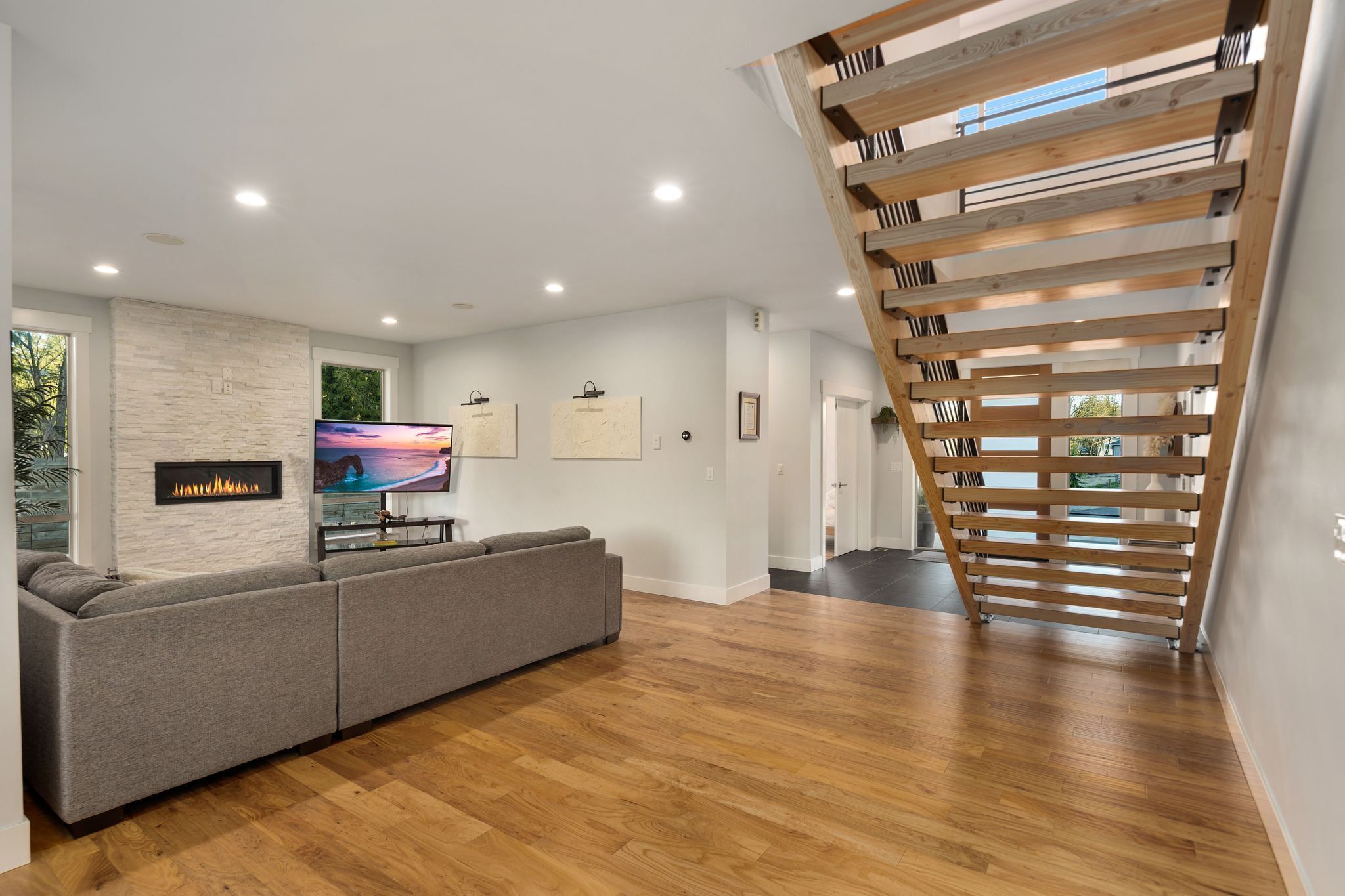 A modern living room with a grey sectional sofa, a flat-screen TV, a fireplace, and a wooden staircase.