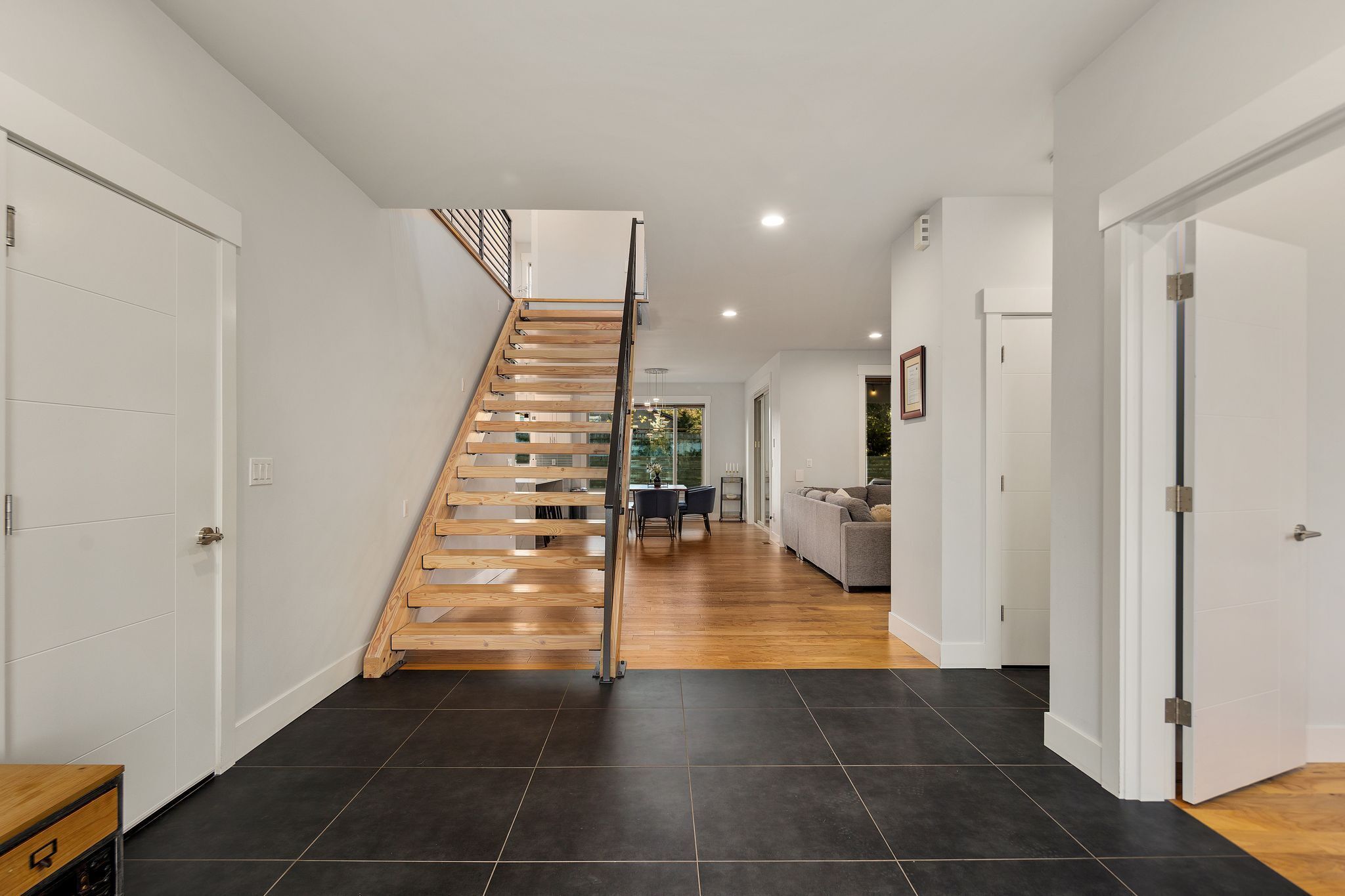 A modern interior with a wooden staircase, black tiled flooring, and a dining area with a table and chairs.