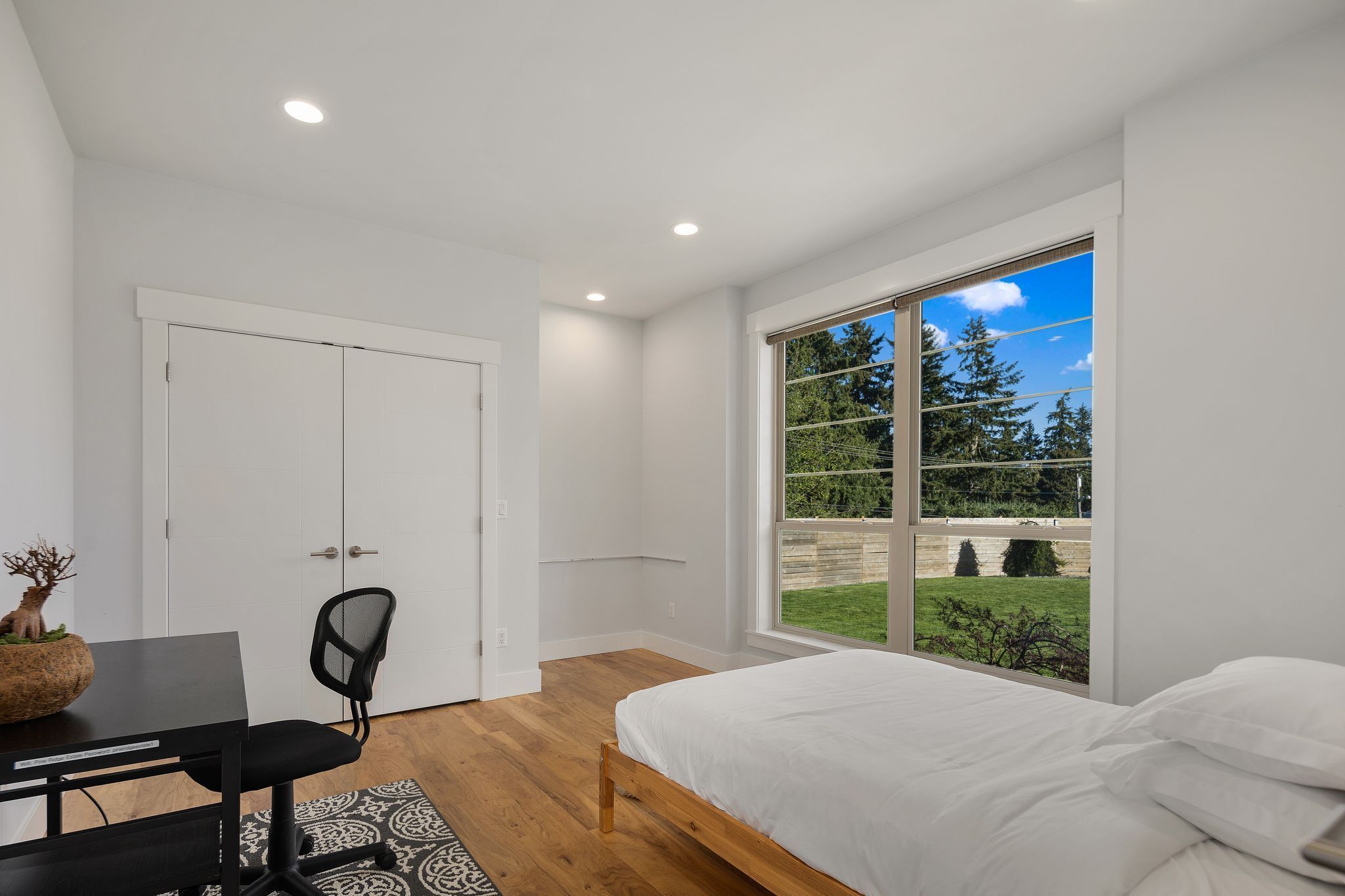 A bedroom with a large window, a bed, a desk with a chair, and a door.