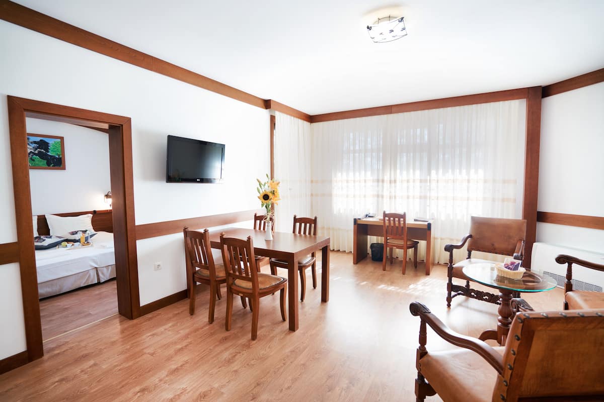 A room with a wooden floor, a dining table with chairs, a TV mounted on the wall, and a bed with a painting.