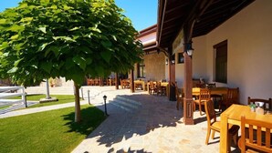 An outdoor seating area with stone flooring, wooden tables and chairs, and a tree providing shade. - Boutique Complex Trakiets (Kaloyanovo)
