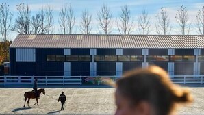 A person in a black top and jeans sitting on a chair with a drink, overlooking an outdoor riding arena with a horse and rider. - Boutique Complex Trakiets (Kaloyanovo)