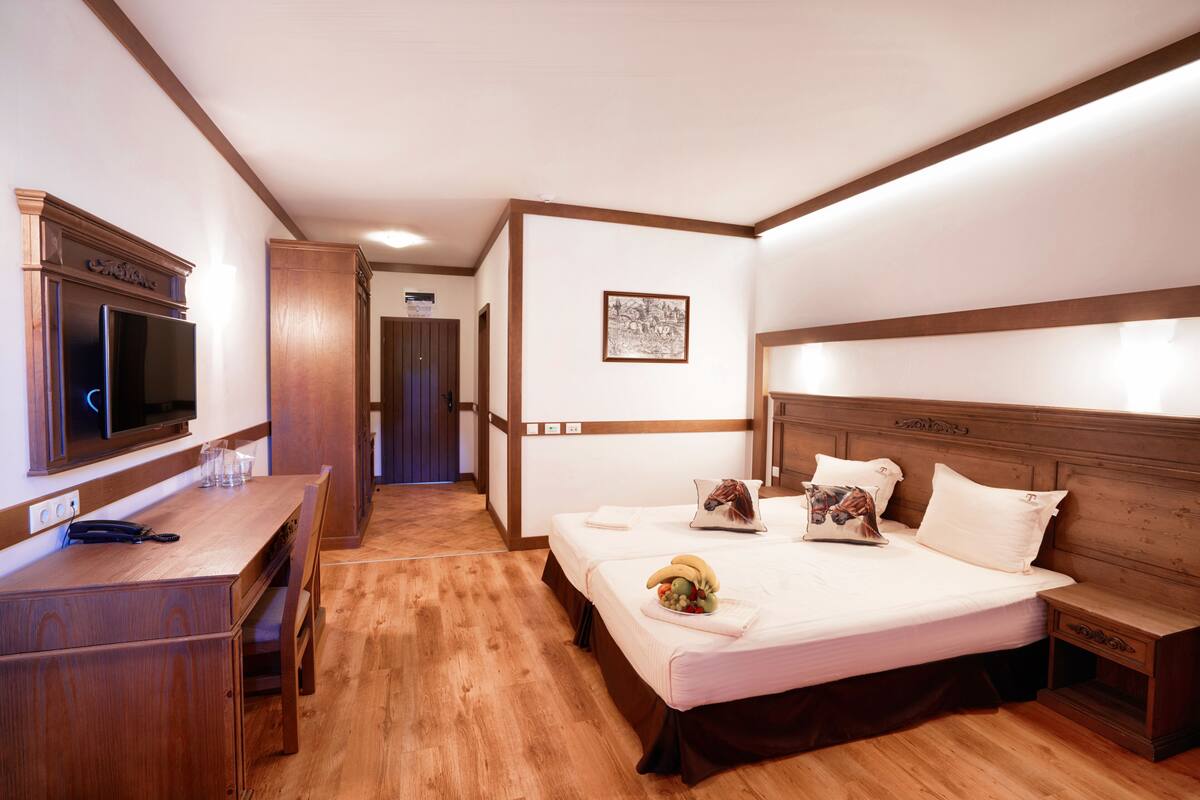 A hotel room with a wooden desk, a bed with pillows and a fruit basket, a television, and a framed picture on the wall.