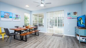 Un coin repas avec une table en bois, des chaises noires et un banc. On y trouve un ventilateur de plafond, une fenêtre avec des stores et une porte surmontée d’une porte blanche.