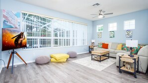 Un salon doté d’une grande fenêtre, d’un ventilateur de plafond, d’un téléviseur posé sur un trépied, d’un canapé, d’une table basse et d’un petit chariot sur lequel se trouve une lampe.