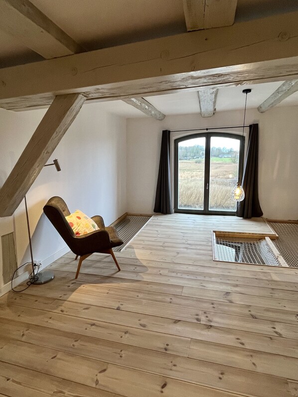 Een ruime zolderkamer met schuine wanden, een houten vloer, een zwarte leren fauteuil, een klein tafeltje en een groot raam met uitzicht op een veld.