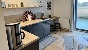 Una cocina moderna con cafetera, estantes con tazas y una planta, un comedor con mesa y sillas, y una ventana con vistas.