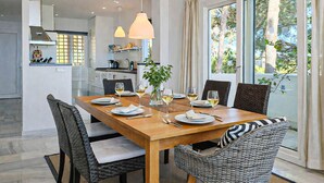 A dining area with a wooden table set for four, surrounded by wicker chairs, a potted plant, and pendant lighting.
