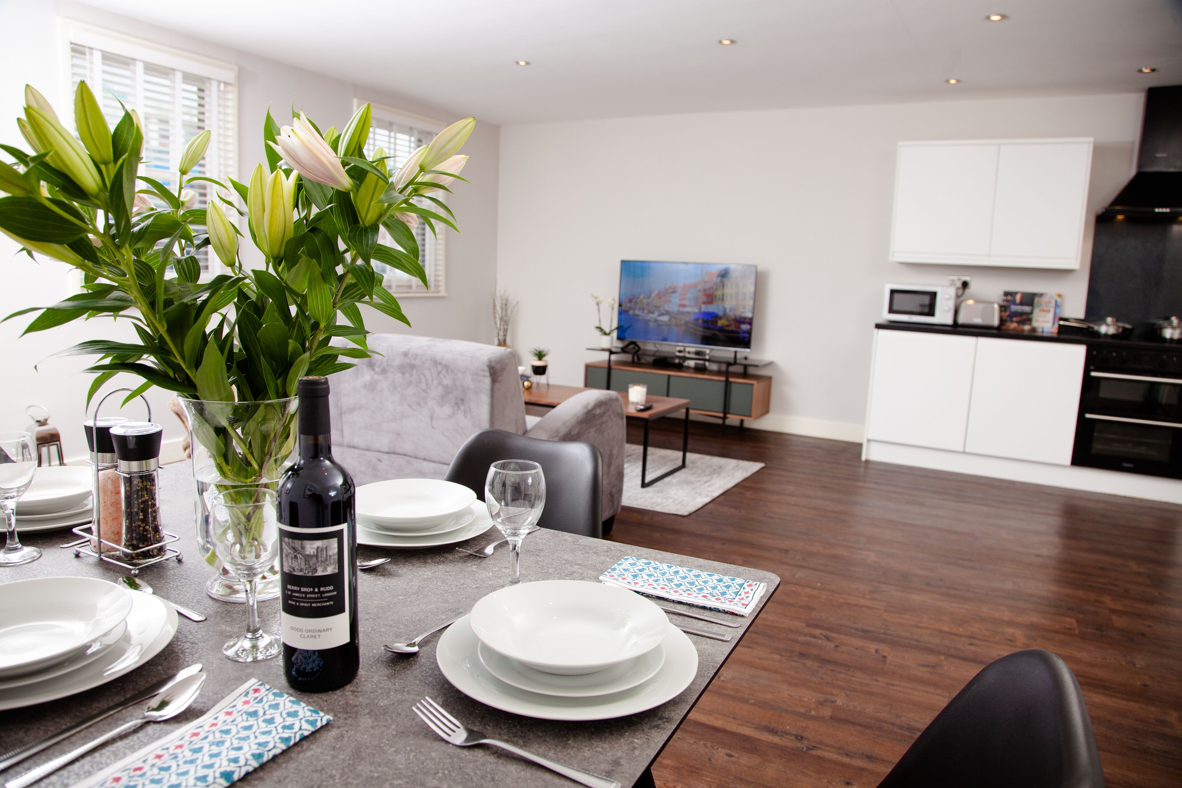 A dining area with a table set for four, a vase with flowers, and a kitchen in the background.