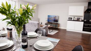 A dining area with a table set for four, a vase with flowers, and a kitchen in the background.