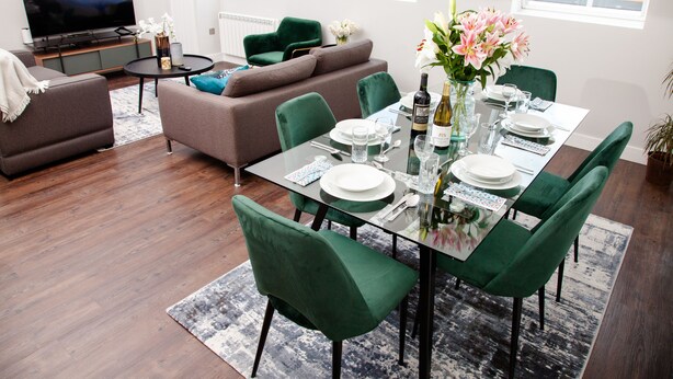 A modern living room with a glass dining table set for six, green upholstered chairs, a brown sofa, a flat-screen TV, and a large window with a view of a building. - High Life Luxury Serviced Apartments  (Swindon)