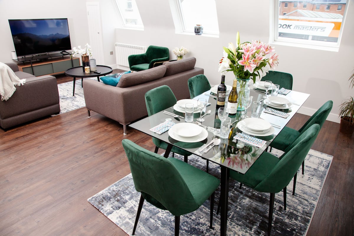 Un salon moderne avec une table à manger en verre pouvant accueillir six personnes, des chaises à revêtement vert, un canapé marron, un téléviseur à écran plat et une grande fenêtre donnant sur un immeuble.