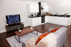 Una sala de estar moderna con un televisor de pantalla plana, una mesa de centro de madera y un sofá gris con cojines naranjas.