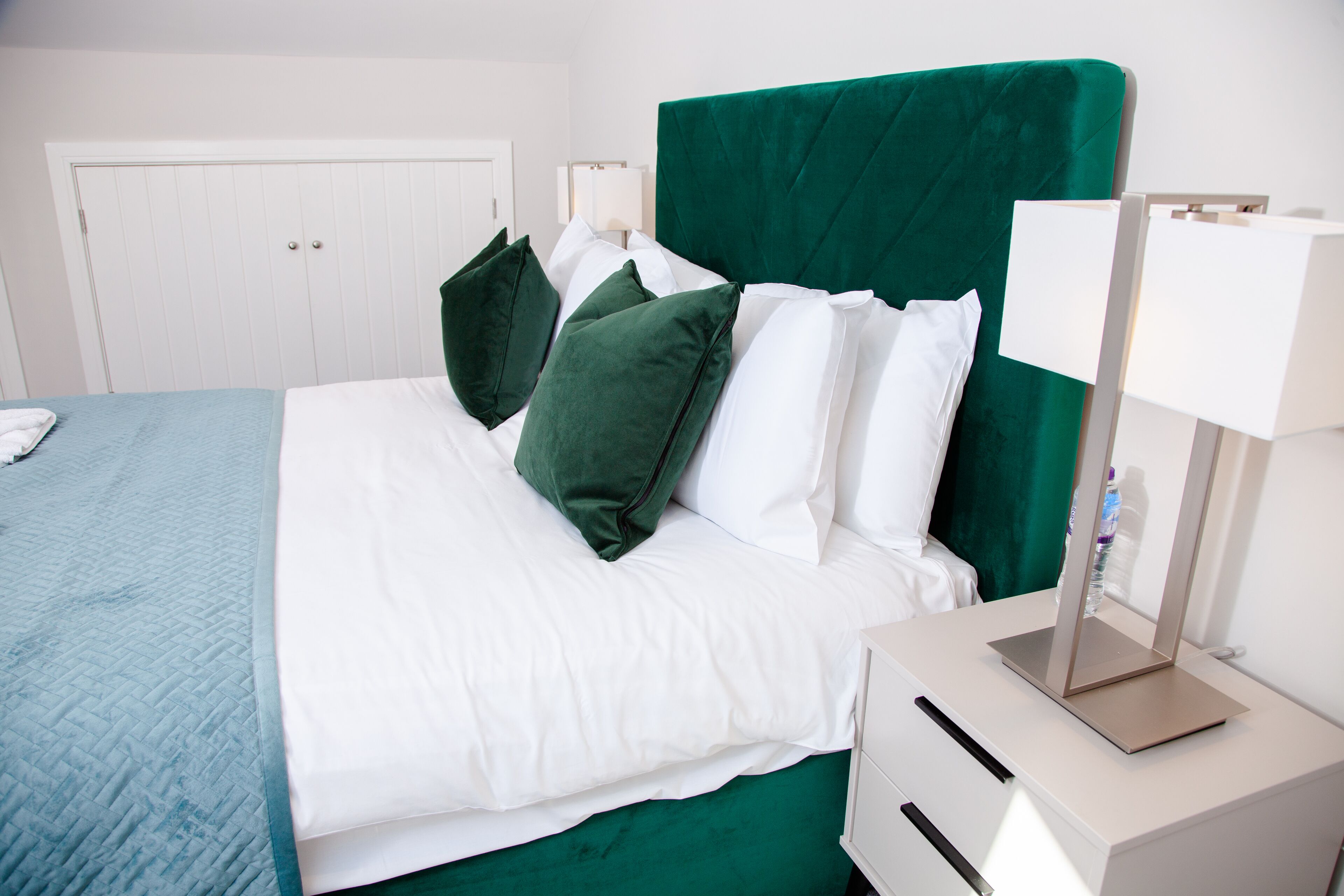 Un letto rifatto con lenzuola bianche e verdi, un comodino con una lampada e un armadio bianco in una camera d'albergo.