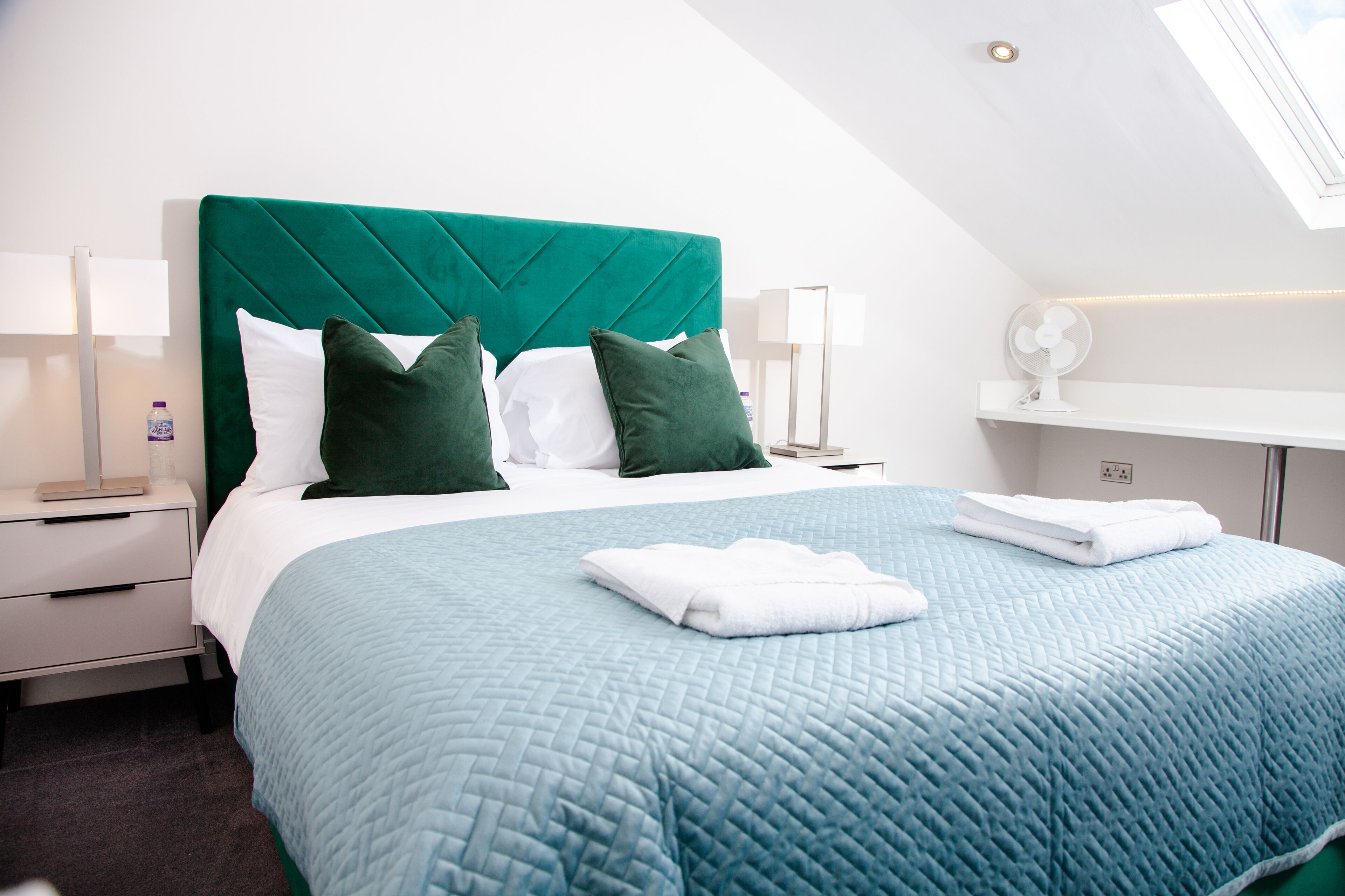 A neatly made bed with a teal quilt, white linens, and green pillows in a modern hotel room with a skylight and a bedside table with a lamp.