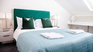 A neatly made bed with a teal quilt, white linens, and green pillows in a modern hotel room with a skylight and a bedside table with a lamp.
