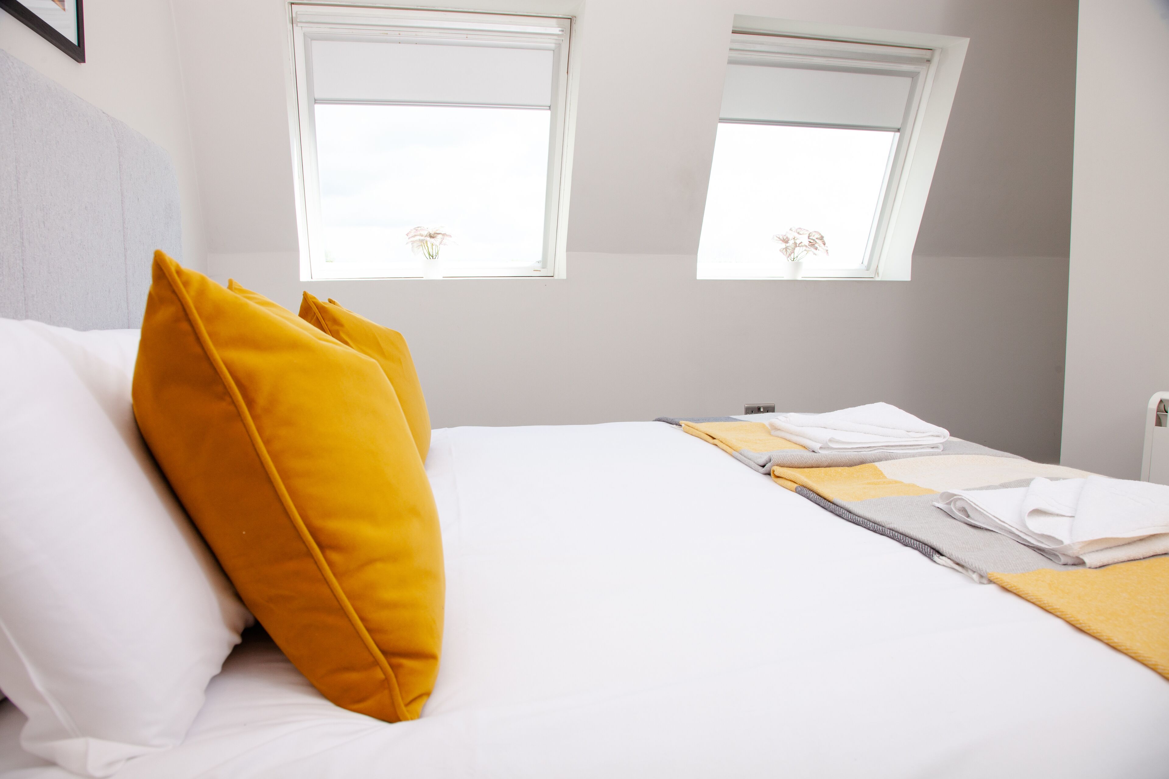 Una cama bien hecha con almohadas blancas y amarillas, una toalla doblada y un pequeño ventanal con vistas a flores.