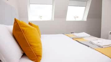 Una cama bien hecha con almohadas blancas y amarillas, una toalla doblada y un pequeño ventanal con vistas a flores.