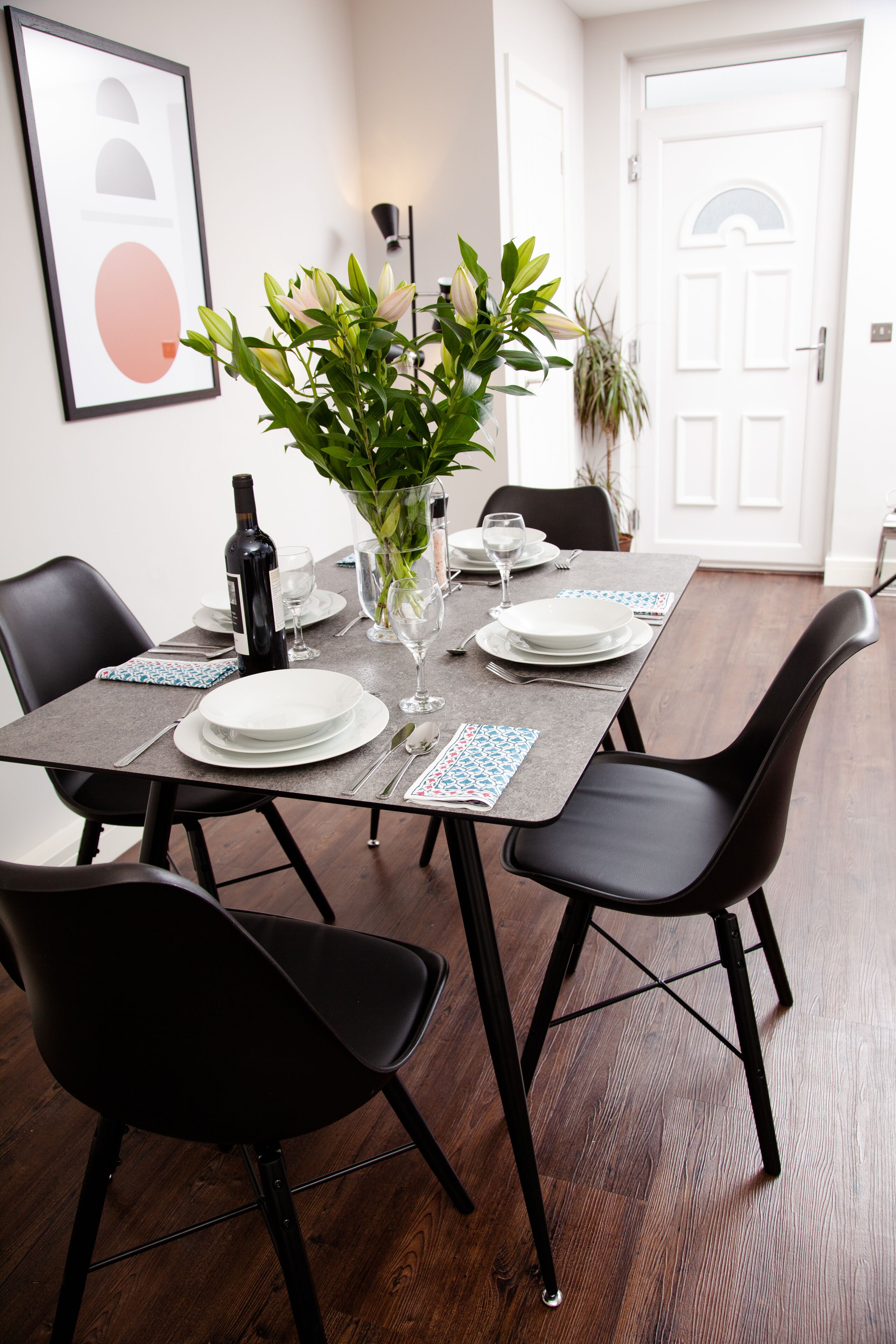 Comedor con una mesa rectangular dispuesta para cuatro personas, rodeada de sillas negras. Un jarrón con follaje verde se coloca sobre la mesa, y se ve una botella de vino. El espacio tiene piso de madera y paredes blancas.