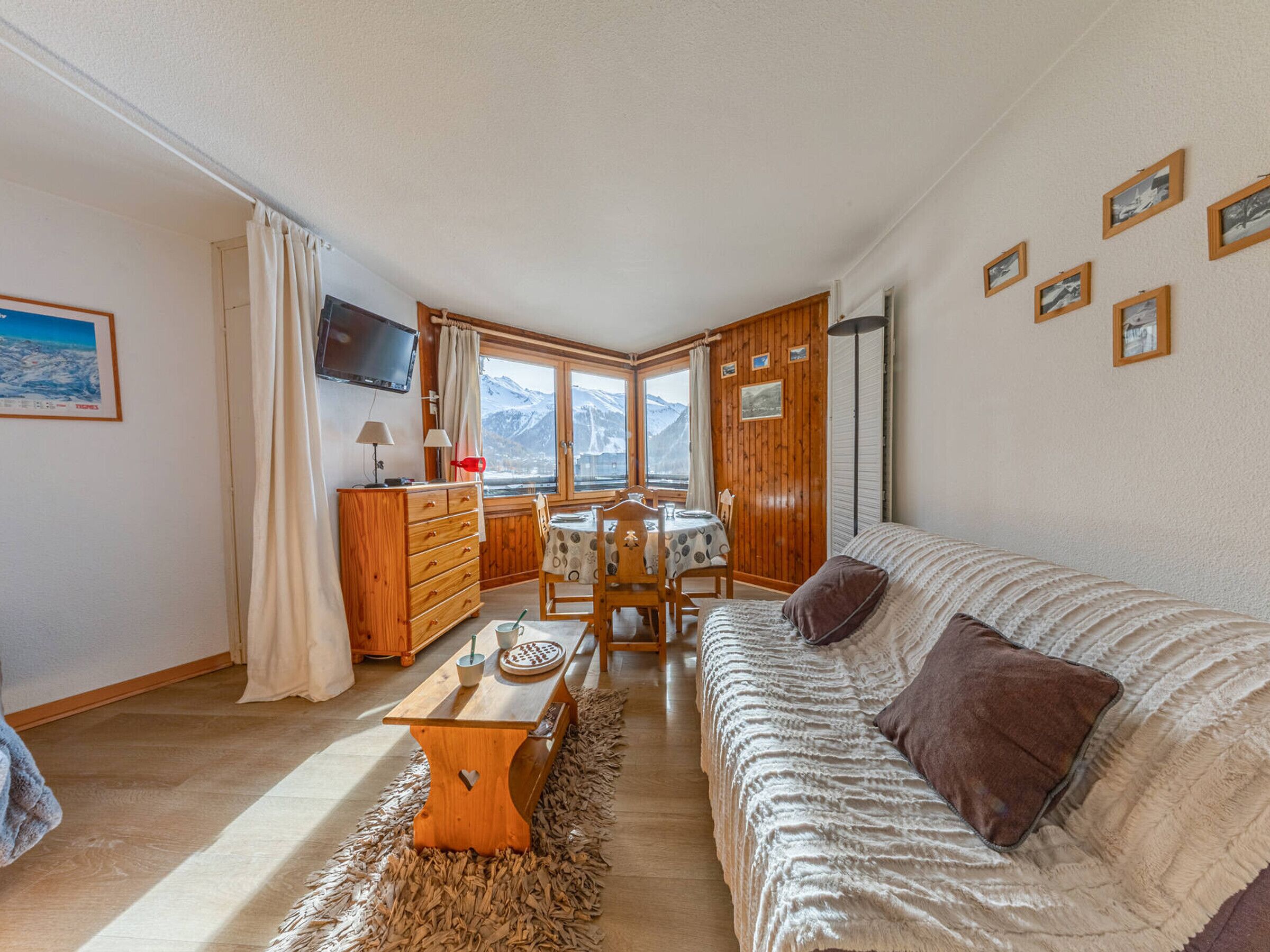 A cozy living room with a wooden sofa, a dining table, and a TV mounted on the wall.