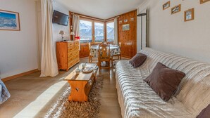 A cozy living room with a wooden sofa, a dining table, and a TV mounted on the wall. - South studio ski-in/ski-out in Val-d'Isère, pets allowed (Val-d'Isère)