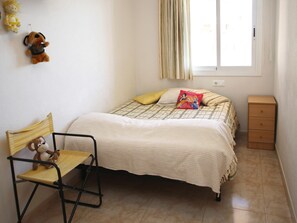 Une chambre à coucher avec un lit, une chaise, une petite commode en bois et une fenêtre avec des rideaux.