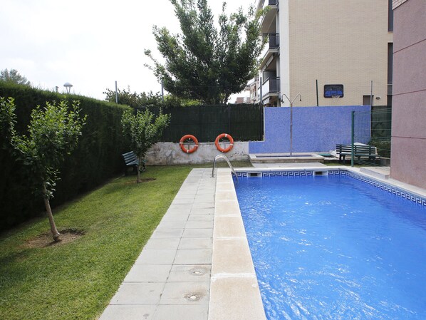 Une piscine bordée de carreaux, une pelouse, un banc et une haie.