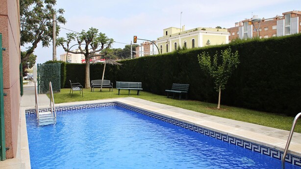 A rectangular swimming pool with a blue mosaic border, surrounded by a low hedge, benches, and a clear sky.
