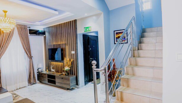 A modern living room with a flat-screen TV mounted on the wall, a wooden entertainment unit, a staircase with a metal railing, and an exit sign. - Grey Top Apartment (Abuja)