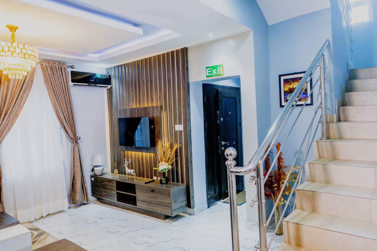 A modern living room with a flat-screen TV mounted on the wall, a wooden entertainment unit, a staircase with a metal railing, and an exit sign.
