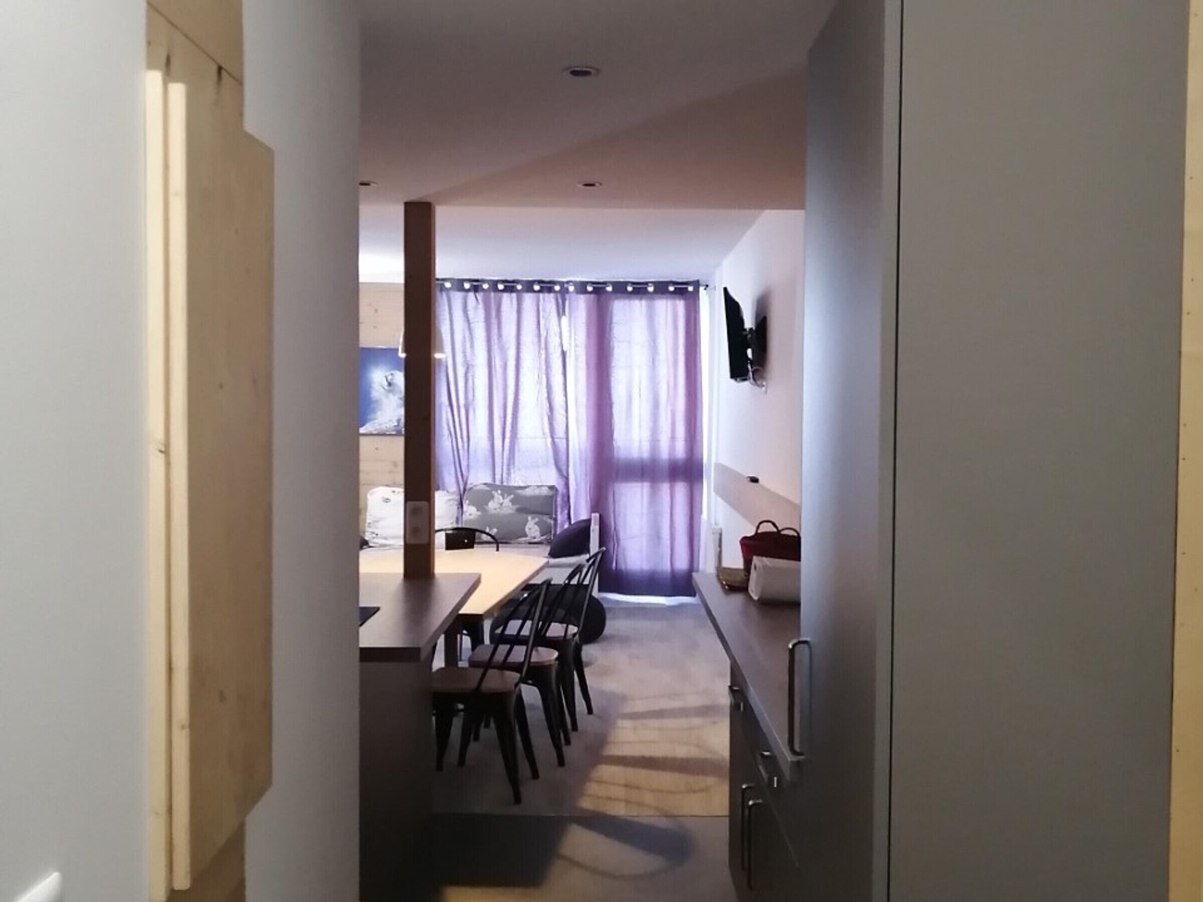 Un estrecho pasillo que da a un comedor con una mesa y sillas, un televisor montado en la pared y una ventana con cortinas.