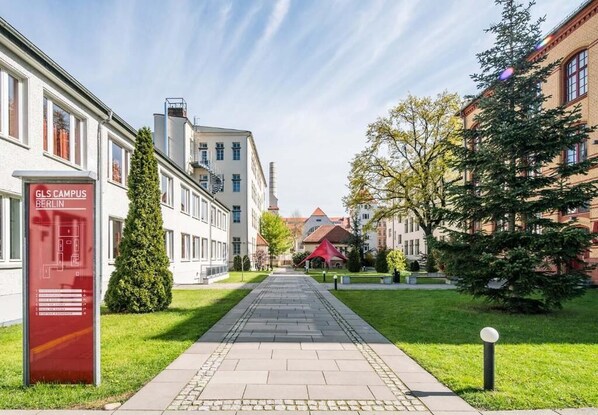 A pathway leading to a building labeled "GLS Campus Berlin" with a red mailbox, surrounded by greenery and traditional architecture.