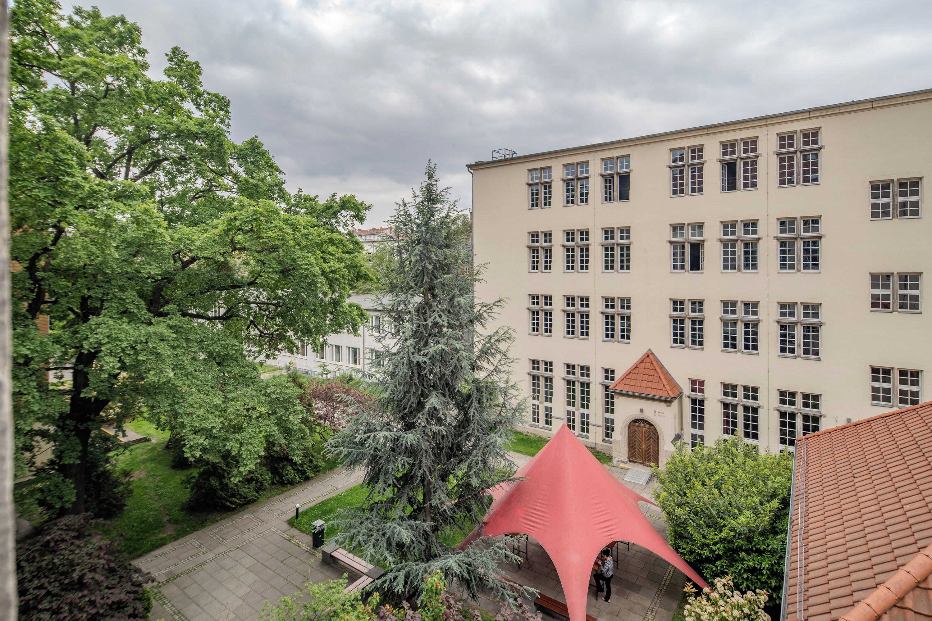 Ein mehrstöckiges Gebäude mit einer symmetrischen Fassade, einem zentralen Eingang mit rotem Dach und einer umliegenden Grünlandschaft mit Bäumen und einem gepflasterten Weg.