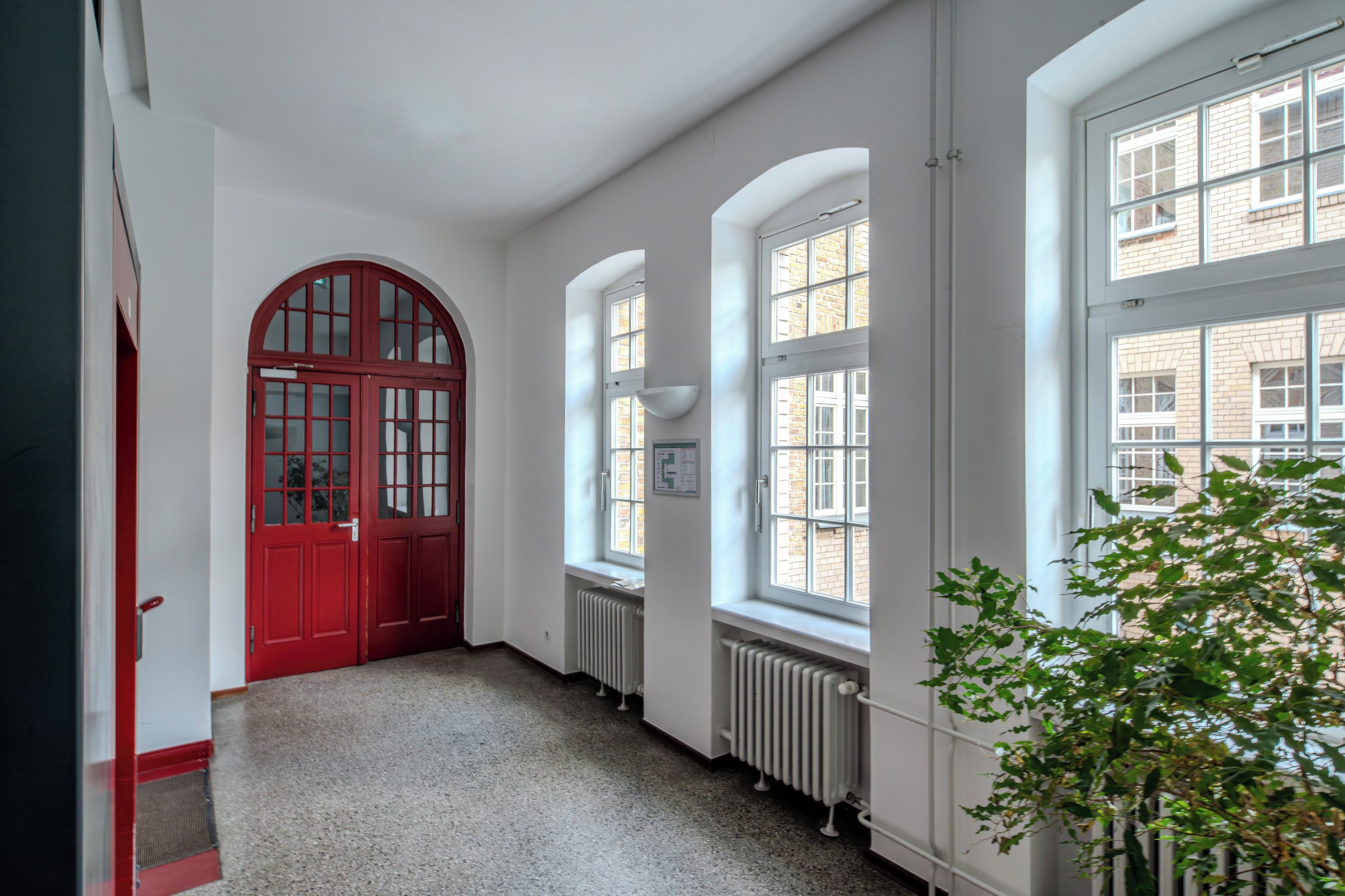 Un couloir avec une porte rouge, des murs blancs et de grandes fenêtres.