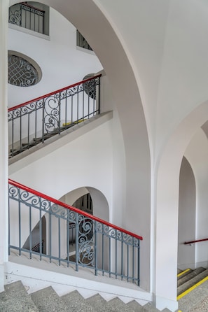 A multi-level interior with white walls and a staircase with ornate metal railings. - GLS Campus Hotel (Berlin)