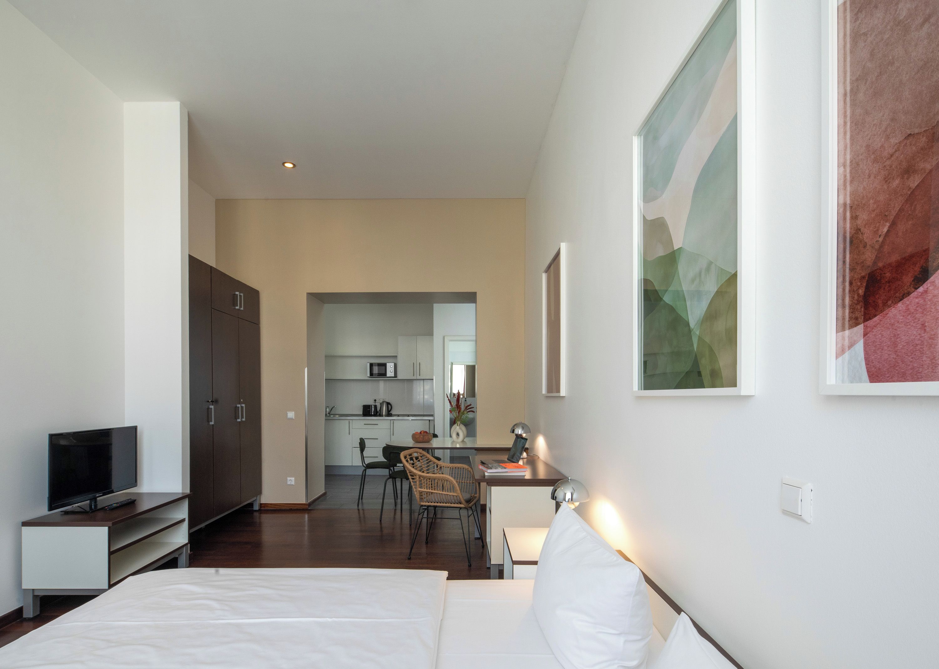 Ein modernes Hotelzimmer mit Bett, Fernseher, Schreibtisch mit Lampe und Blick auf die Küche und den Essbereich.