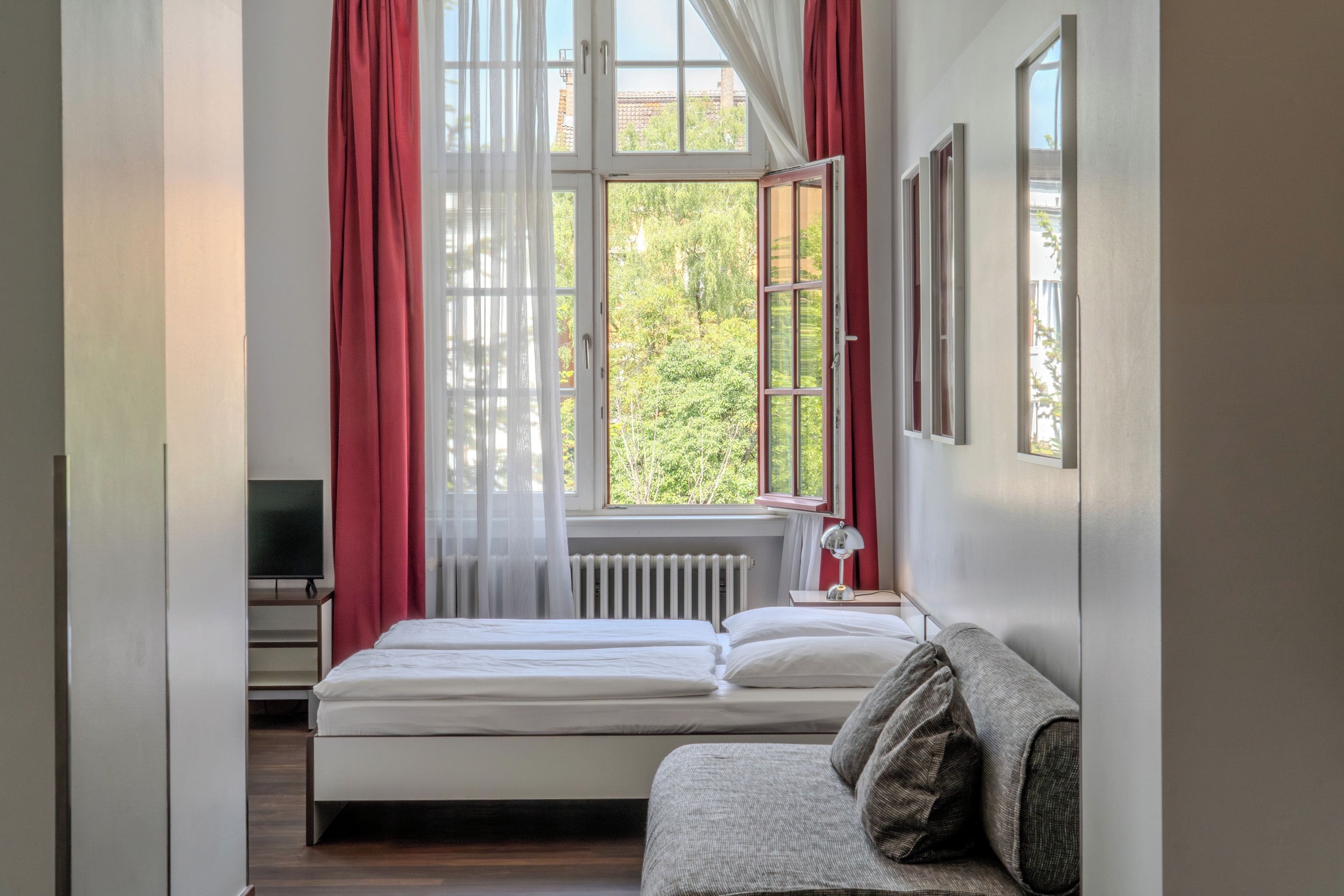 Ein Schlafzimmer mit einem Bett, einem Nachttisch mit Lampe, einem Fernseher und einem Fenster mit roten Vorhängen.