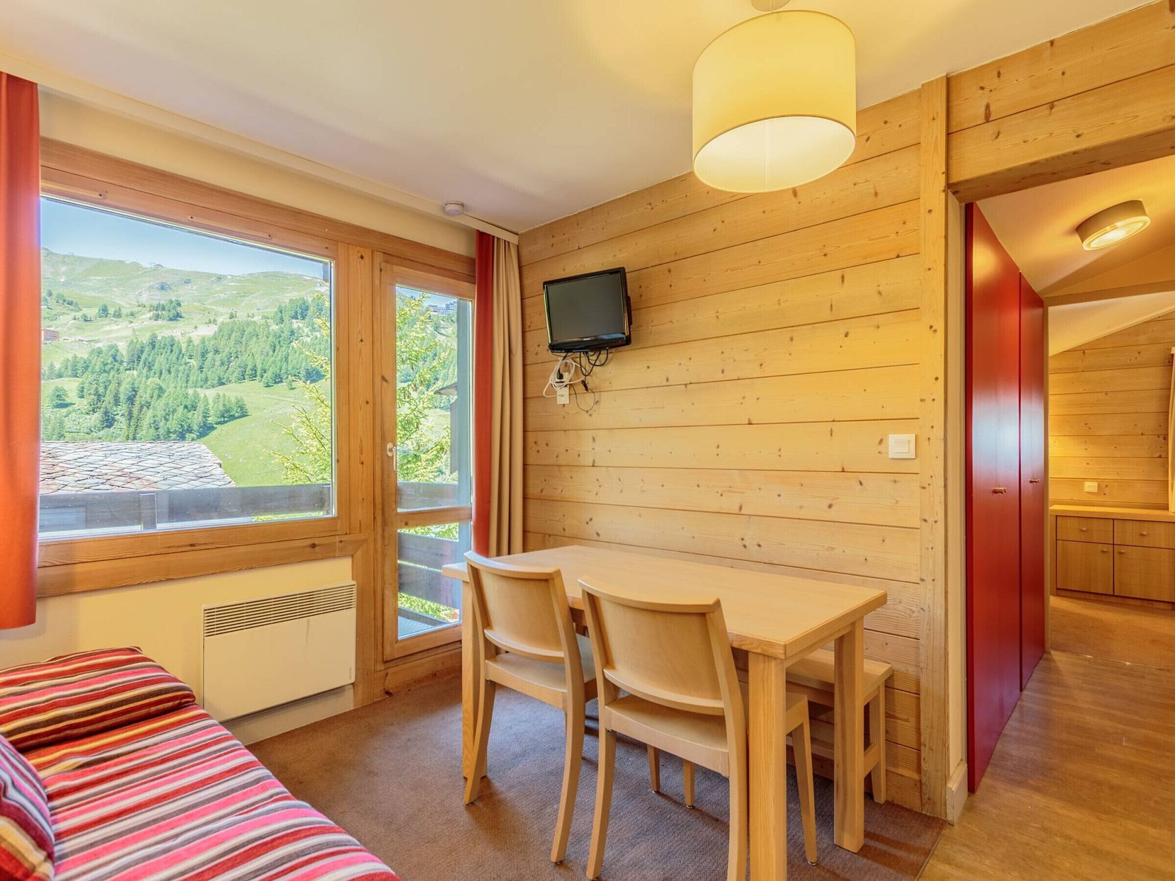 A cozy room with a wooden interior, a dining table with chairs, a TV mounted on the wall, and a large window offering a scenic view.