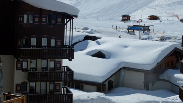Un pueblo cubierto de nieve en la montaña, con un edificio de madera que tiene balcones y un telesilla al fondo.