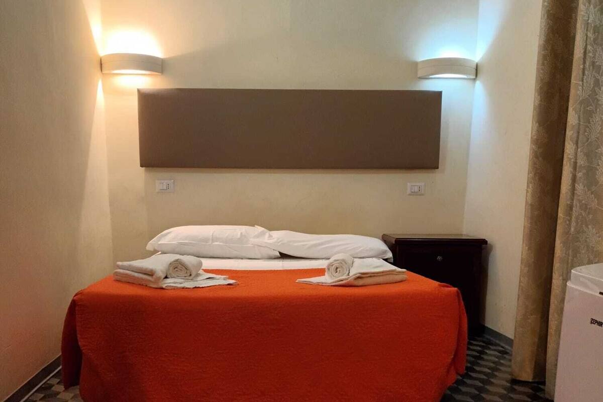 A hotel room with a checkered floor, a bed with an orange bedspread, white pillows, and a dark nightstand.