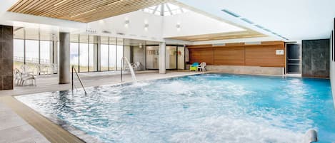 Una piscina interior con techo de madera, paredes de cristal transparente y vistas al mar.